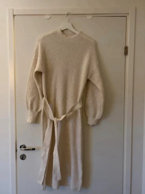 Beige stickad långklänning från ONLY - Mysig beige stickad långklänning från ONLY med rund hals, långa ärmar och knytskärp i midjan. Perfekt för dig som gillar en avslappnad men ändå stilren look. Använd 1 gång 