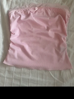 Ljusrosa ribbad spets tubtopp - Supersöt ljusrosa tubtopp med ribbad struktur och spetskant upptill. Toppen har en liten rosett framtill och är axelbandslös, perfekt för varma dagar eller lager på lager. Mjuk och stretchig känsla.