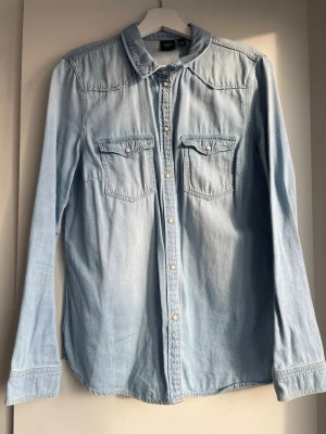 Ljusblå jeansskjorta med knappar - Säljer en ljusblå jeansskjorta från vero moda  med klassisk krage, tryckknappar framtill och två bröstfickor med lock. Skjortan har lång ärm och är tillverkad i mjuk bomull, perfekt för en avslappnad och trendig look.