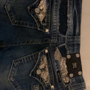 Miss Me jeans med paljettdetaljer - Snygga mörkblå jeans från Miss Me med glittriga paljettdetaljer och dekorativa knappar på bakfickorna. Jeansen har klassisk femficksdesign, låg midja och bootcut. Ett par defekter därav priset. Mått: midja - 37cm innerbenet - 80cm