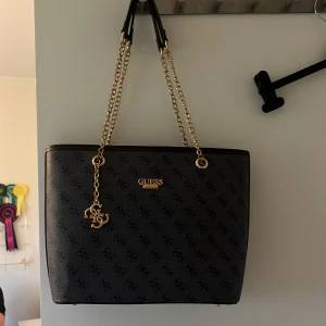 Svart axelväska från Guess med kedja - Säljer en svart axelväska från Guess med guldfärgade kedjehandtag och logomönster över hela väskan. Väskan har dragkedja upptill, en ficka med dragkedja på baksidan och Guess-logga i guld framtill. Insidan är fodrad med Guess-tryck. ⚠️Fake⚠️