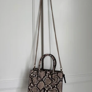 Liten väska med ormskinnsmönster - Liten väska från HM med ormskinnsmönster. Väskan har en guldkedja som även går att haka av om man bara vill använda lilla handtaget. Väskan har dragkedja och även ett litet fack i. Skriv för mer info och bilder.