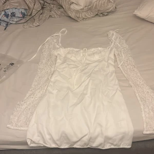 Vit kort klänning med spetsärmar - Supersöt vit kort klänning med långa spetsärmar och knytband vid axlarna. Klänningen har en smickrande v-ringning och fina spetsdetaljer upptill. Tillverkad i mjuk polyesterblandning som känns skön mot huden. Perfekt för dig som vill ha en romantisk och trendig look.