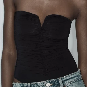 Svart bandeau tubtopp med V-ringning - Säljer en svart bandeau tubtopp med snygg V-ringning framtill och lätt rynkade detaljer över hela toppen. Toppen är tight och axelbandslös, perfekt att styla med jeans eller kjol för en cool look.