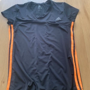 Svart Adidas t-shirt med orange ränder - Sportig svart t-shirt från Adidas med korta ärmar och två orange ränder längs sidorna. T-shirten har v-ringning och är tillverkad i ett lätt och funktionellt material som passar perfekt för träning. Adidas-logga på bröstet.