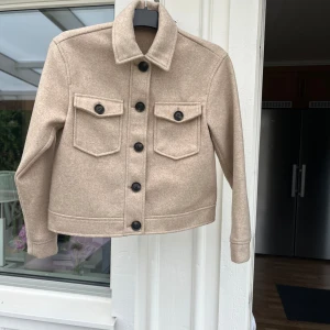 Beige jacka  - Stilren beige jacka från Vero Moda med stora svarta knappar och två bröstfickor med lock. Jackan har klassisk krage och raka ärmar. Materialet känns mjukt och har en ullig look som ger en trendig vibe.
