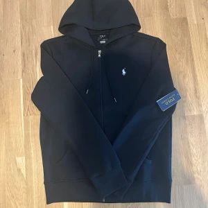 Svart hoodie från Polo Ralph Lauren - Svart hoodie från Polo Ralph Lauren med dragkedja och klassisk huva. Tröjan har det ikoniska broderade logotypen på bröstet och är tillverkad i mjuk bomull. Perfekt för en chill och stilren look.