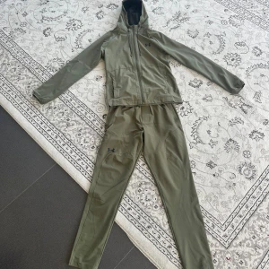 Under Armour grön träningsset / kostym - Snyggt olivgrönt träningsset från Under Armour med jacka och byxa. Jackan har huva, dragkedja och meshfoder, medan byxorna har justerbar midja och diskret logga på benet. Perfekt för dig som gillar sportig stil och funktionella plagg.