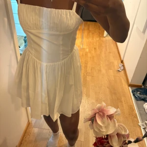 Vit kort sommarklänning med axelband - Har aldrig använt den och prislappen är fortfarande på .Supersöt vit kort klänning med smala axelband och rynkad överdel. Kjolen är luftig och har mycket vidd, perfekt för varma dagar. 