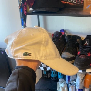 Vit keps från Lacoste i bomull - Snygg vit keps från Lacoste med klassisk krokodillogga broderad på sidan. Justerbar rem baktill med Lacoste-märke. Tillverkad i 100% bomull, perfekt för dig som gillar stilrena accessoarer och vill ha en clean look.