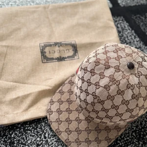 Gucci beige keps med GG-mönster - Säljer en snygg beige keps från Gucci med klassiskt brunt GG-monogram över hela kepsen. Den har en böjd skärm och justerbar rem bak. På sidan finns den ikoniska gröna och röda randen. Materialet är bomull och polyester, perfekt för dig som vill ha en lyxig streetstyle.