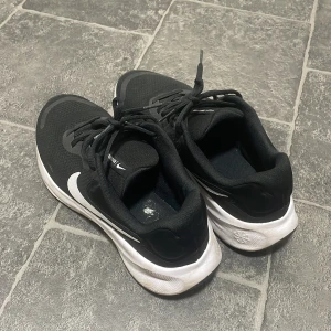 Svarta Nike sneakers med vit sula - Svarta Nike sneakers med vit sula och stor vit swoosh på sidan. Ovandelen är i mesh för bra andningsförmåga och skorna har klassisk snörning. Perfekta för träning eller vardagsstil. Sulan är mönstrad för extra grepp och komfort.