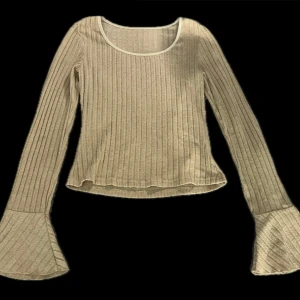 Beige ribbad topp med trumpetärmar - Superfin beige ribbad topp med långa trumpetärmar och rund halsringning. Toppen har en figurnära passform och är tillverkad i ett mjukt material som känns skönt mot huden. Perfekt för dig som gillar en stilren men ändå unik look.
