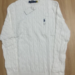 Vit kabelstickad tröja Polo Ralph Lauren - Vit kabelstickad tröja från Polo Ralph Lauren med klassisk blå logga broderad på bröstet. Tröjan har rund halsringning och långa ärmar, perfekt för en clean och stilren look. Tillverkad i mjuk bomull för extra komfort.