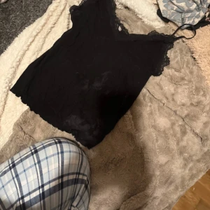Svart spetskantad linnetopp - Säljer en svart linnetopp med tunna axelband och v-ringning. Toppen har fina spetsdetaljer längs halsringningen och är tillverkad i ett mjukt, lätt material. Perfekt att styla med jeans eller kjol för en trendig look.