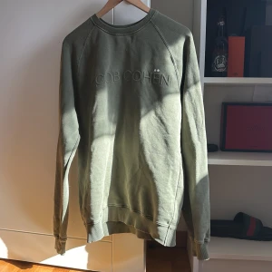 Olivgrön sweatshirt från Jacob Cohën - Olivgrön sweatshirt från Jacob Cohën med broderad logga framtill. Tröjan har rund hals, långa ärmar och ribbade muddar vid ärmslut och nederkant. Tillverkad i mjuk bomull, perfekt för en chill och stilren look. Tröjan är väl använd och är lite urtvättad. Kan finnas små defekter och därför är priset som det är  passar l/m