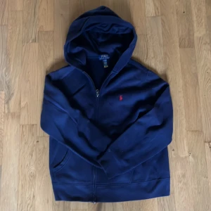 Marinblå hoodie från Polo Ralph Lauren - Snygg marinblå hoodie från Polo Ralph Lauren med dragkedja och huva. Tröjan har det klassiska röda Polo-logot broderat på bröstet och är tillverkad i mjuk bomull. Perfekt för chill dagar och har praktiska fickor framtill.