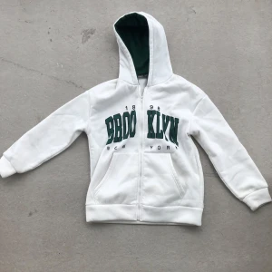 Vit hoodie med grön text från SHEIN - Vit hoodie med dragkedja från SHEIN, stor grön text 'BROOKLYN' framtill och mörkgrönt foder i huvan. Klassisk college-stil med känguruficka och långa ärmar. Perfekt för chill dagar eller när du vill ha en sportig vibe.