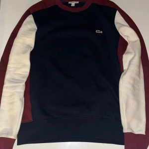Marinblå sweatshirt från Lacoste - Snygg marinblå sweatshirt från Lacoste med kontrastärmar i vinrött och vitt. Klassisk rund hals och broderad krokodil-logga på bröstet. Tröjan har långärmad design och är gjord i mjuk bomull för skön komfort. Perfekt för chill dagar och streetwear.
