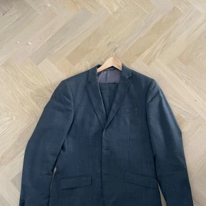 Mörkblå kavaj från Riley  - Snygg mörkgrå kavaj med klassisk krage och två fickor framtill. Kavajen har en stilren look och är perfekt för dig som vill ha en tidlös stil. Passar lika bra till skjorta som t-shirt. Fodrad insida för extra komfort. Storlek 152. Kommer med byxorna.