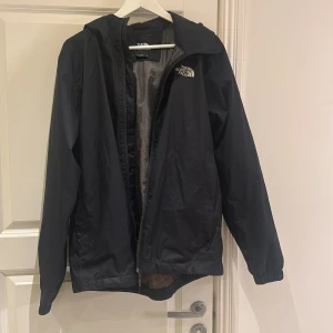 Svart vindjacka från The North Face - Svart vindjacka från The North Face med huva och dragkedja framtill. Jackan har meshfoder på insidan, elastiska muddar vid ärmsluten och logga på bröstet. Perfekt för blåsiga dagar och enkel att slänga på sig över en hoodie.