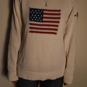 Vit stickad med USA-flagga - Vit stickad tröja med broderad amerikansk flagga på bröstet och blå text på ärmen. Tröjan har ribbade muddar och lång ärm 