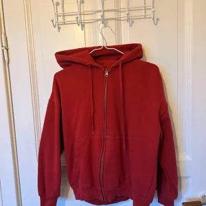 Röd hoodie med dragkedja från Junkyard - Röd hoodie från Junkyard i storlek S. Klassisk modell med huva, dragsko och hel dragkedja framtill. Mjukt material och känguruficka framtill. Perfekt för chill dagar eller när du vill ha en enkel streetstyle-look.