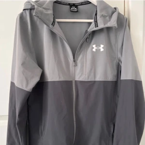 Grå vindjacka från Under Armour - Snygg tvåfärgad grå vindjacka från Under Armour med huva och dragkedja hela vägen. Jackan har en vit logga på bröstet, justerbar huva och praktiska fickor. Perfekt för dig som gillar sportig och clean stil. Väldigt bra skick
