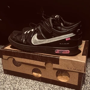 Nike Dunk Low Off-White svart/silver - Nike Dunk Low Off-White i svart skinn och textil med silvrig Swoosh, vita och svarta snören samt unika Off-White-detaljer och text på sidan. Platt sula och rosa accent på mellansulan. Riktigt hypad sneaker med streetkänsla.