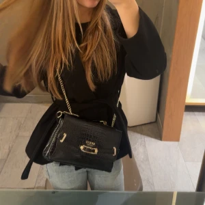 Svart axelväska från Guess med kedja - Säljer en svart axelväska från Guess med croco-mönstrad yta och snygga guldfärgade detaljer. Väskan har en kedjerem som ger en lyxig känsla och ett stilrent metallspänne framtill. Perfekt storlek för det viktigaste och passar till många outfits. Nypris 1700kr