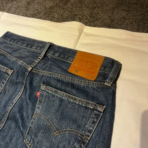 Levi's 501 jeans blå W33 L34 - Klassiska Levi's 501 jeans i blå denim med raka ben och normal passform. Byxorna har fem fickor, knappgylf och den ikoniska läderpatchen bak i midjan. Snygga kontrastsömmar och röd Levi's-tagg på bakfickan. Perfekta till en avslappnad streetstil.