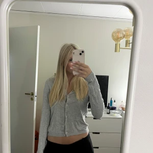 Grå kofta från lager 157 - Säljer en stilren grå croppad kofta med långa ärmar och knappar framtill. Tröjan har en v-ringad hals och är gjord i mjukt material som känns skönt mot huden. Perfekt för dig som gillar en enkel och clean look.