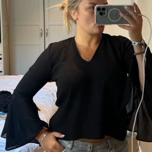 Svart blus med trumpetärm från H&M Divided - Svart blus med v-ringning och vida trumpetärmar från H&M Divided. Toppen har öppen rygg med snygg knyt-detalj bak. Materialet är lätt och lite transparent, perfekt för att ge en cool vibe till din outfit.