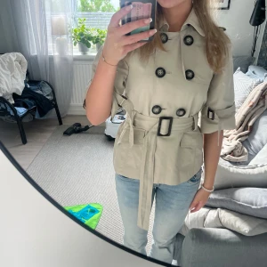 Trenchcoat  - Snygg beige trenchcoat med korta ärmar, stora svarta knappar och bälte i midjan. Jackan har klassisk krage, fickor framtill och detaljerade spännen vid ärmarna. Perfekt för dig som vill ha en stilren och trendig look till våren eller hösten.