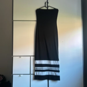 Svart axelbandslös långklänning - Snygg svart axelbandslös långklänning med raka linjer och vita ränder nertill. Klänningen har en enkel och stilren design som ger en cool vibe. Materialet ser ut att vara mjukt och följsamt, perfekt för dig som gillar minimalistisk stil.