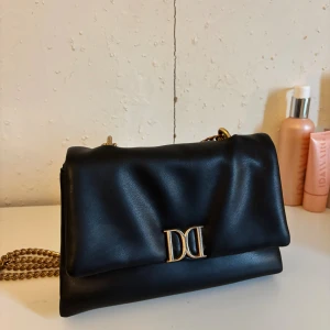 Don Donna väska - Snygg svart axelväska från Dolce & Gabbana i mjukt skinn med guldfärgad kedjerem och ikonisk DD-logga framtill. Perfekt storlek för det viktigaste och ger en lyxig touch till din outfit.