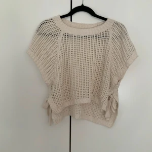 Beige stickad topp med knyt i sidan - Säljer en beige stickad topp med korta ärmar och snyggt hålmönster. Toppen har bred ribbad kant vid halsen och coola knytband i sidorna som ger extra detalj. Perfekt för lager-på-lager och chill stil.