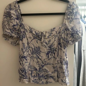 Blommig blus med puffärm från H&M - Supersöt blommig blus från H&M i vitt och blått med puffärmar och smockad rygg. Toppen har knyt fram och volangdetaljer vid ärmslut. Perfekt för dig som gillar romantisk och trendig stil.
