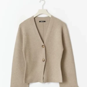 Beige kofta med knappar från Gina Tricot - Stilren beige kofta från Gina Tricot med v-ringning och två snygga knappar framtill. Koftan har lång ärm och är tillverkad i mjukt material som passar perfekt för lager-på-lager. Enkel att matcha med både jeans och kjol.