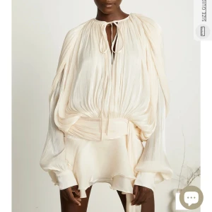 Champagnefärgad klänning miss rosier - Superdrömmig kortklänning i champagnefärgad sidenmix. Klänningen har voluminösa långärmar, och flowy volangkjol. Plisserad struktur och knytning vid halsen ger en lyxig och unik vibe. Perfekt för dig som vill sticka ut med en statement piece. Aldrig använd förutom provning, blev aldrig av köpte den i vintras för sommaren. Jag är storlek small och skulle säga att klänningen är XS/Small.