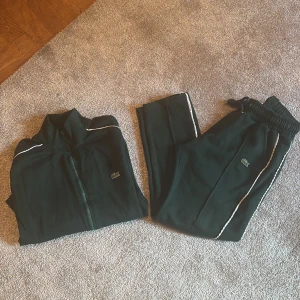 Mörkgrön zip-tröja plus mjukisbyxor från Lacoste - Mörkgrön ziptröja & mjukisbyxor från Lacoste med dragkedja framtill och vit piping längs axlarna. Klassisk krokodil-logga på bröstet. Sportig look med hög krage och mjukt material, perfekt för en chill och stilren outfit.