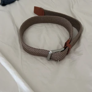 Beige flätat bälte med silverfärgat spänne - Snyggt beige flätat bälte med silverfärgat metallspänne och bruna skinn-detaljer vid ändarna. Perfekt accessoar för att lyfta din outfit med en stilren touch. Bältet är flexibelt och passar till många olika looks.