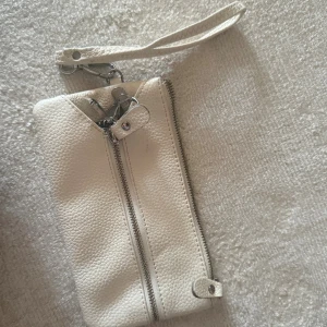 Vit kuvertväska i skinn med dragkedja - Snygg vit kuvertväska i präglat skinn med silverfärgade detaljer och praktisk handledsrem. Väskan har två dragkedjor framtill och en smart nyckelring på insidan. Perfekt för att hålla ordning på småsaker och nycklar.