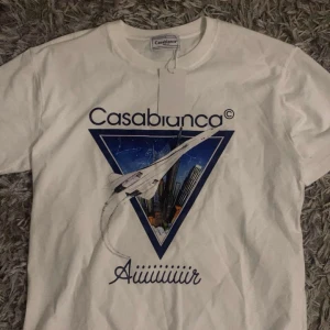 Vit Casablanca t-shirt med tryck - Casablanca t-shirt i vit bomull med stort grafiskt tryck framtill som visar ett flygplan och stadssilhuett i blått och vitt. Klassisk rund hals och korta ärmar. Snygg logga och texten 'Aiiiiiiiir' under motivet. Perfekt för dig som gillar streetwear och coola prints.