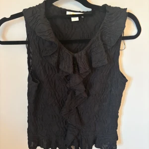 Svart volangblus från Gina Tricot - Supersnygg svart blus från Gina Tricot med volangdetaljer längs v-ringningen och framtill. Blusen är ärmlös och har ett lätt transparent, vågigt tyg som ger en cool struktur. Den är bara använd en gång och är i väldigt bra skick. Den är i storlek 170.💓