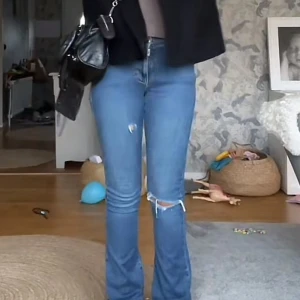 Blå bootcut lågmidjade jeans med slitningar - Snygga lågmidjade blå jeans med bootcut passform och slitningar på knäet och låret. Jeansen har klassisk femficksdesign och stängs med dragkedja och knapp. Perfekta för en avslappnad och trendig look.