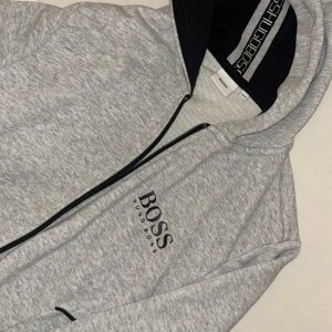 Grå hoodie från Hugo Boss med dragkedja - Snygg ljusgrå hoodie från Hugo Boss med svart logga på bröstet. Tröjan har hel dragkedja, huva med svart innerfoder och två fickor framtill. Perfekt för en chill och sportig look. Tillverkad i mjuk bomullsblandning för extra komfort.