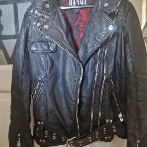 Svart skinnjacka Jofama by Kenza - Säljer en ikonisk svart skinnjacka från Jofama by Kenza. Jackan har coola silvriga dragkedjor, bälte med nitar och klassisk bikerstil. Fodret är rött med svart spetsmönster för extra edge. Skinnjackan sitter ganska tajt på kroppen att jag skulle säga XXS-XS som storlek. Skriv för fler bilder🙏