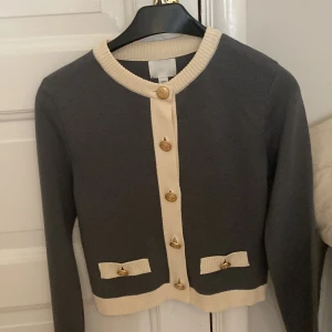Grå kofta med gulddetaljer från H&M - Supersnygg grå kofta från H&M i storlek XS med krämvita ribbade kanter och guldknappar framtill. Koftan har två dekorativa fickor och långärmad passform. Perfekt för dig som gillar klassisk men trendig stil.