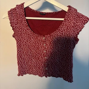 Röd blommig topp från Brandy Melville - Supersöt röd topp från Brandy Melville med små vita blommor, knappar framtill och lätt croppad passform. Toppen har korta ärmar med vågig kant. Passar storlek XXS-M